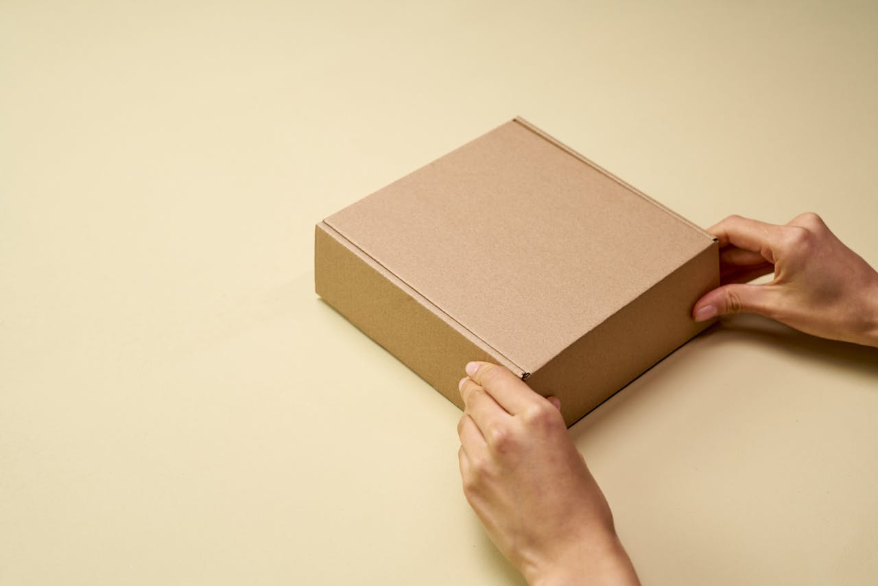 our-services-2 A close-up of hands opening a plain brown cardboard box on a beige surface, perfect for themes of shipping or packaging.